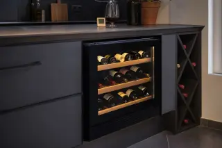 Compact electric wine cellar under a kitchen counter with wine bottles neatly organised, anti-UV glass door, digital hygrometer nearby, and a glass of red wine on the countertop in a modern urban kitchen.