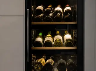 Compact multi-zone electric wine cellar built into a modern kitchen, with glass door, digital panel, and neatly arranged wine bottles visible in different temperature compartments.