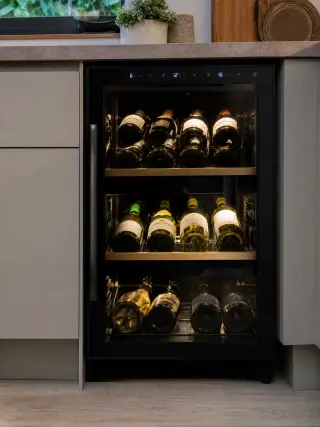 Compact multi-zone electric wine cellar built into a modern kitchen, with glass door, digital panel, and neatly arranged wine bottles visible in different temperature compartments.