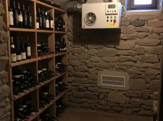 Rustic wine cellar with stone walls, wooden racks of wine bottles, a mechanical ventilator near the ceiling, and a passive ventilation grille by the floor, softly lit by natural light.